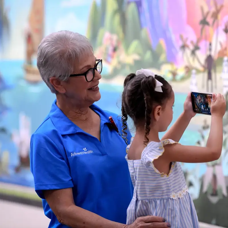An older woman in a blue AdventHealth shirt helps a young girl take a photo of a colorful mural.
