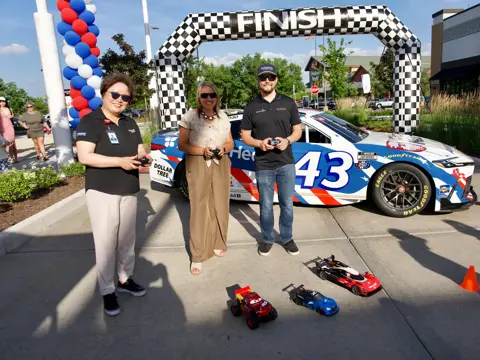 Three people hold remote controls while standing in front of a branded race car beneath a checkered finish arch, with small remote-controlled cars on the ground.