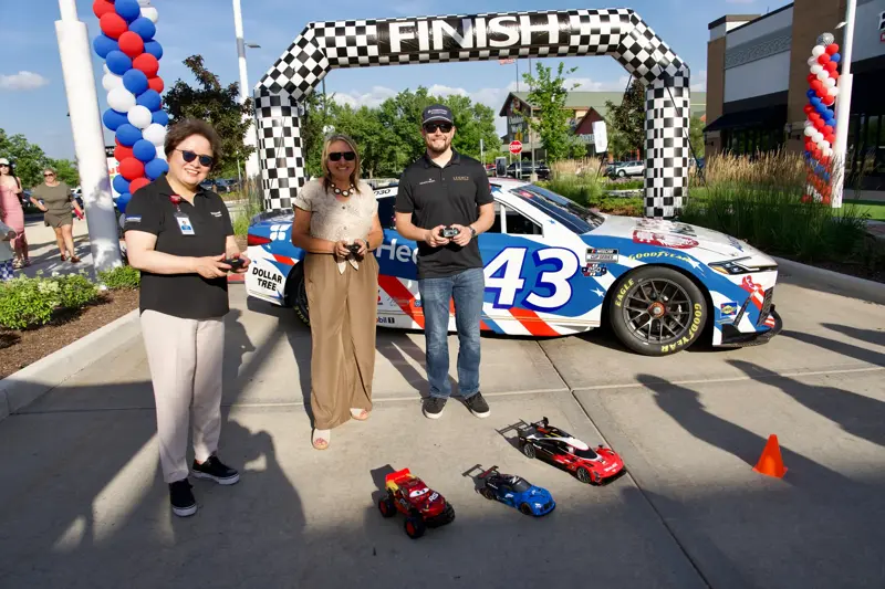 Three people hold remote controls while standing in front of a branded race car beneath a checkered finish arch, with small remote-controlled cars on the ground.