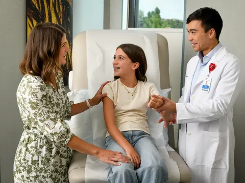 A young girl with braces sits in a chair as a doctor and a woman stand beside her, smiling.
