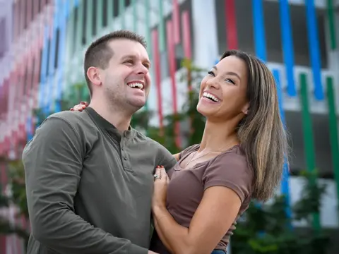 A smiling couple stands close together in front of a colorful building, embodying warmth and connection.