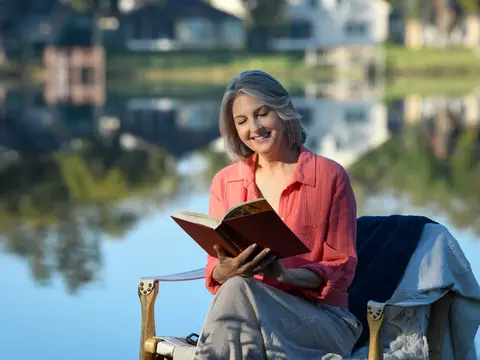 A smiling woman in a pink shirt reads a book while sitting on a chair by a lake.