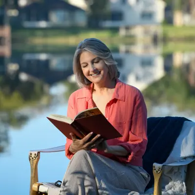 A smiling woman in a pink shirt reads a book while sitting on a chair by a lake.