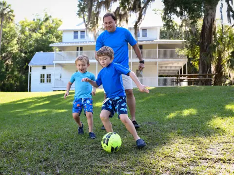 Father and two young sons playing soccer in the yard of a white house.