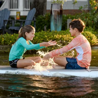 Two children sitting on a paddleboard, splashing water and smiling.