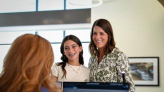 A smiling woman and a young girl with braces stand behind a woman looking at a computer screen.