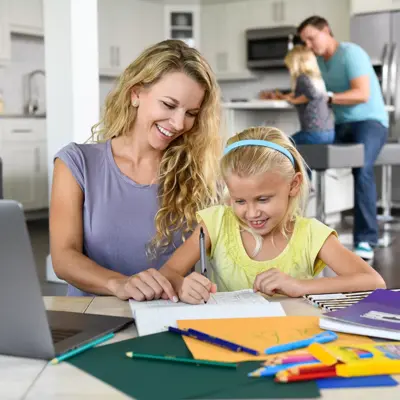 Mother and daughter working on homework at a kitchen table with a laptop and art supplies.