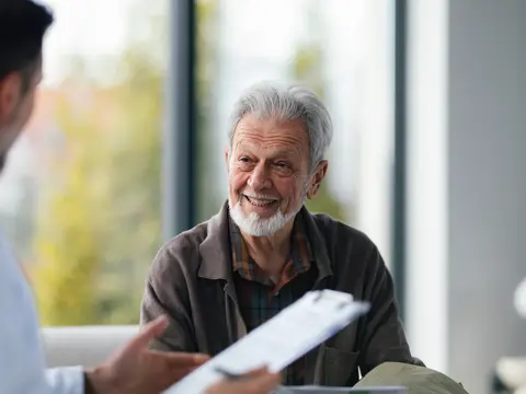Senior man with gray hair and beard smiling warmly while engaged in conversation with a health care provider in a bright, modern clinic room with large windows