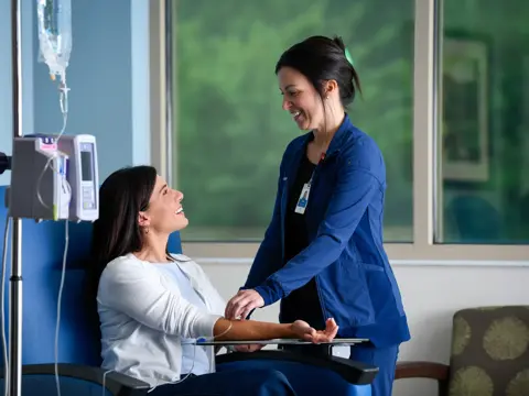 Health care professional in blue scrubs attending to a smiling patient seated in a blue chair receiving IV treatment in a bright medical room