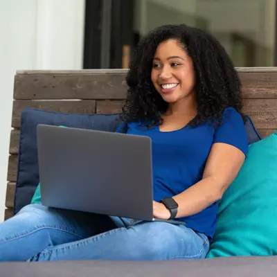 A smiling woman in a blue shirt sits on a wooden bench with a laptop.