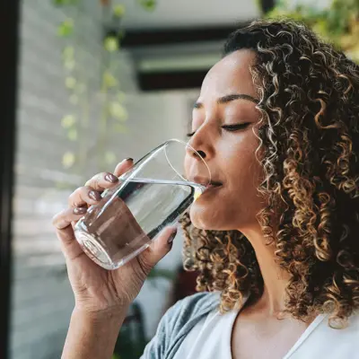 Woman with curly hair drinking a glass of water while seated indoors near a window
