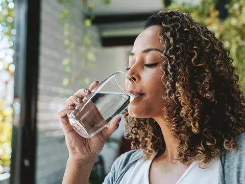 Woman with curly hair drinking a glass of water while seated indoors near a window