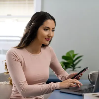 A woman in a pink shirt uses her phone while sitting at a desk with a laptop and candle.