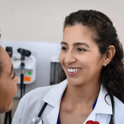 A smiling doctor in a white coat with a stethoscope around her neck talks to a patient.