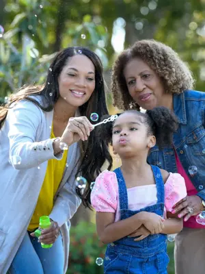 Three generations of women smiling and blowing bubbles in a park.