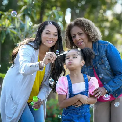 Three generations of women smiling and blowing bubbles in a park.
