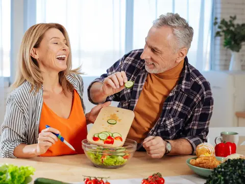 Older couple in a bright kitchen preparing a fresh salad together, with the man playfully holding a cucumber slice while the woman laughs