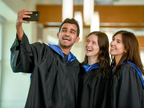 Three graduates in black and blue robes smiling and taking a selfie in a hallway.