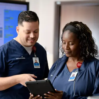 Two AdventHealth employees in scrubs, smiling and looking at a tablet in front of a large screen.