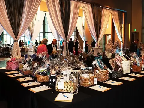 Holiday charity ball silent auction display featuring gift baskets, bid sheets and festive decorations in an elegant event venue with draped curtains and formal attendees.