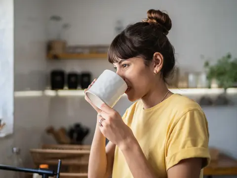 A woman in a yellow shirt drinks from a white mug while standing in a bright kitchen with plants and shelves in the background