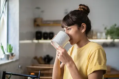 A woman in a yellow shirt drinks from a white mug while standing in a bright kitchen with plants and shelves in the background
