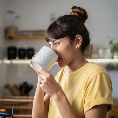 A woman in a yellow shirt drinks from a white mug while standing in a bright kitchen with plants and shelves in the background