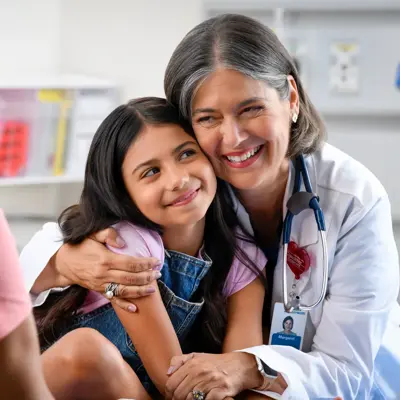 A smiling doctor with a stethoscope around her neck hugs a young girl, while another woman watches.