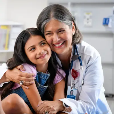 A smiling doctor with a stethoscope around her neck hugs a young girl, while another woman watches.