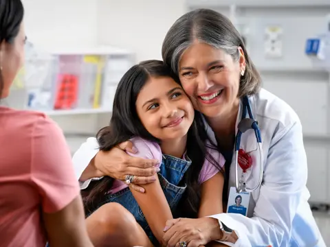 A smiling doctor with a stethoscope around her neck hugs a young girl, while another woman watches.