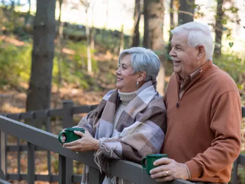 Elderly couple smiling and holding green mugs while standing on a wooden deck in a forest.
