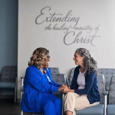 Two women sitting in chairs facing each other, holding hands and smiling in a waiting room with text on the wall reading 'Extending the healing ministry of Christ'