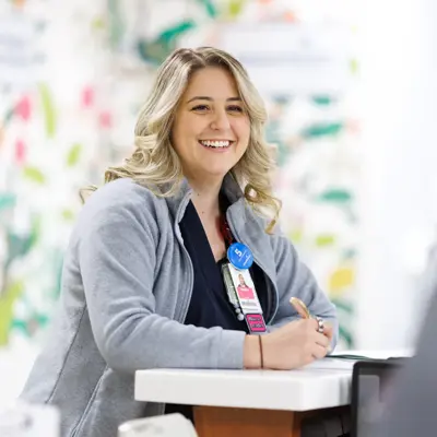 Smiling nurse in gray cardigan and navy scrubs sitting at a desk with a computer in a bright hospital setting