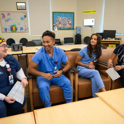 Four healthcare professionals in scrubs, smiling and conversing in a hospital room.