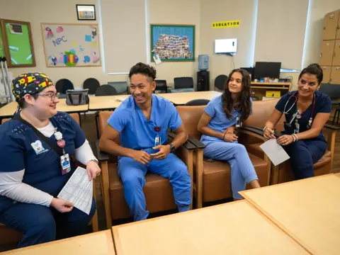 Four healthcare professionals in scrubs, smiling and conversing in a hospital room.