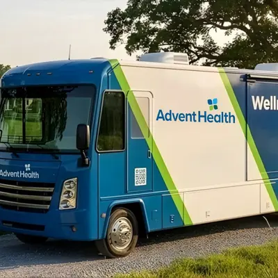 Blue and white AdventHealth mobile clinic parked on gravel near a barn, with wellness message on the side.