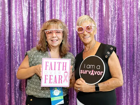 Two women wearing festive pink glasses and holding breast cancer awareness signs in front of a purple backdrop.