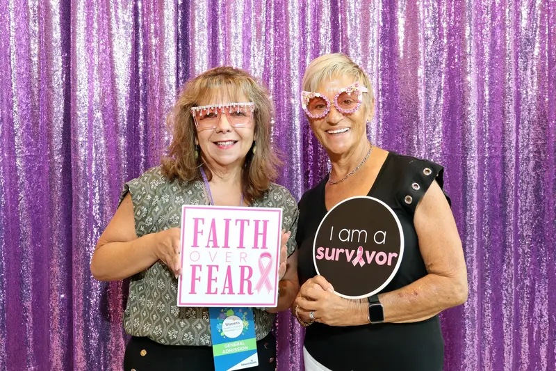 Two women wearing festive pink glasses and holding breast cancer awareness signs in front of a purple backdrop.