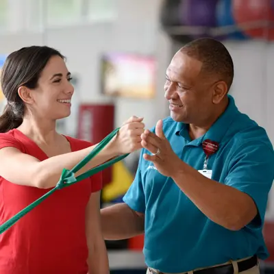A smiling woman in a red shirt is assisted by a man in a blue shirt with a resistance band.