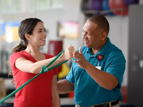 A smiling woman in a red shirt is assisted by a man in a blue shirt with a resistance band.