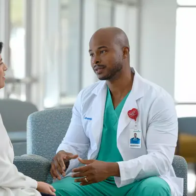 A doctor in a white coat and green scrubs talks to a patient in a white shirt in a hospital waiting room.