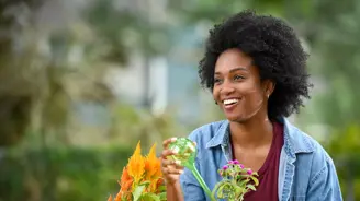 A smiling woman in a denim jacket and green gloves watering plants in a garden.