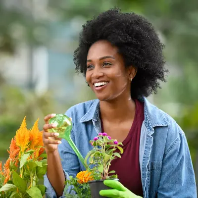 A smiling woman in a denim jacket and green gloves watering plants in a garden.