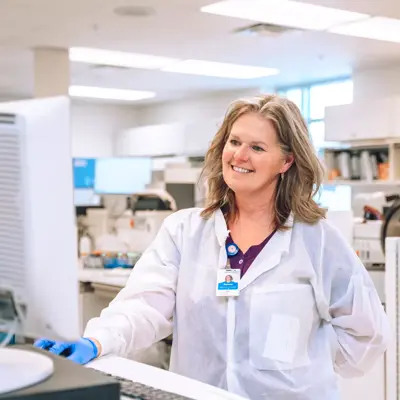 A smiling woman in a lab coat stands at a computer in a lab setting.