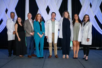 Eight people pose together on a stage with white draped decorations, including several wearing white coats, smiling at the camera.