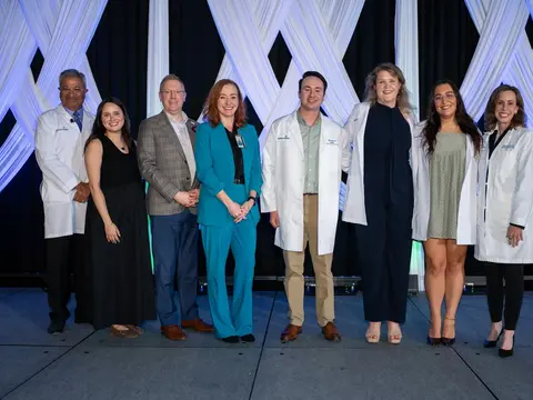 Eight people pose together on a stage with white draped decorations, including several wearing white coats, smiling at the camera.