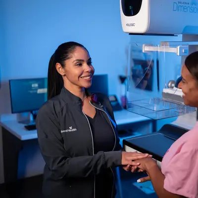 AdventHealth team member in black uniform smiling and holding hands with a patient in a pink top standing beside a Hologic Selenia Dimensions mammography machine in a blue-lit imaging room