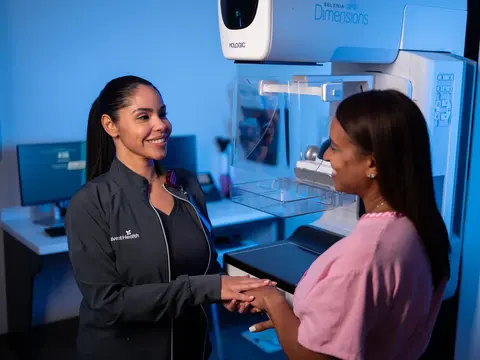 AdventHealth team member in black uniform smiling and holding hands with a patient in a pink top standing beside a Hologic Selenia Dimensions mammography machine in a blue-lit imaging room