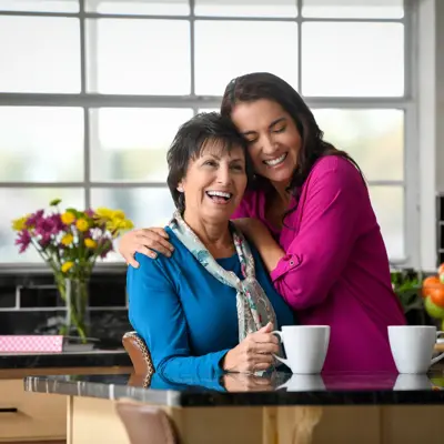 Two women hugging in a kitchen with coffee cups and fresh fruit on the counter.