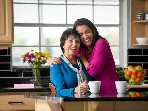 Two women hugging in a kitchen with coffee cups and fresh fruit on the counter.